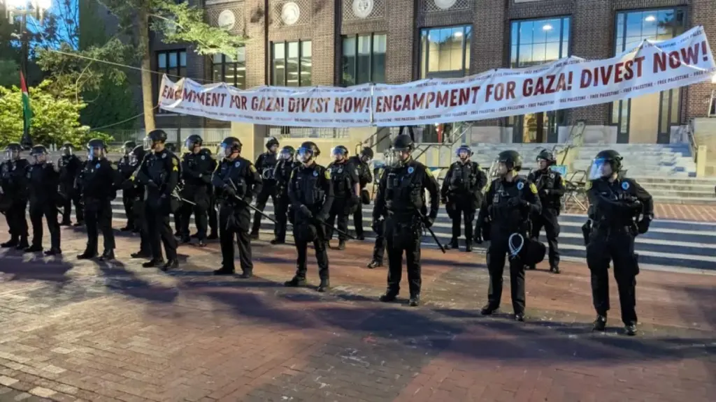 Police in riot gear stand in front of the sign that reads "Encampment for Gaza! Divest Now!"